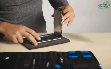 تعمیر کار در حال تعمیر گوشی باز شده با رنگ نقره ای بر روی میز چوبی کنار کیف وسایل اش